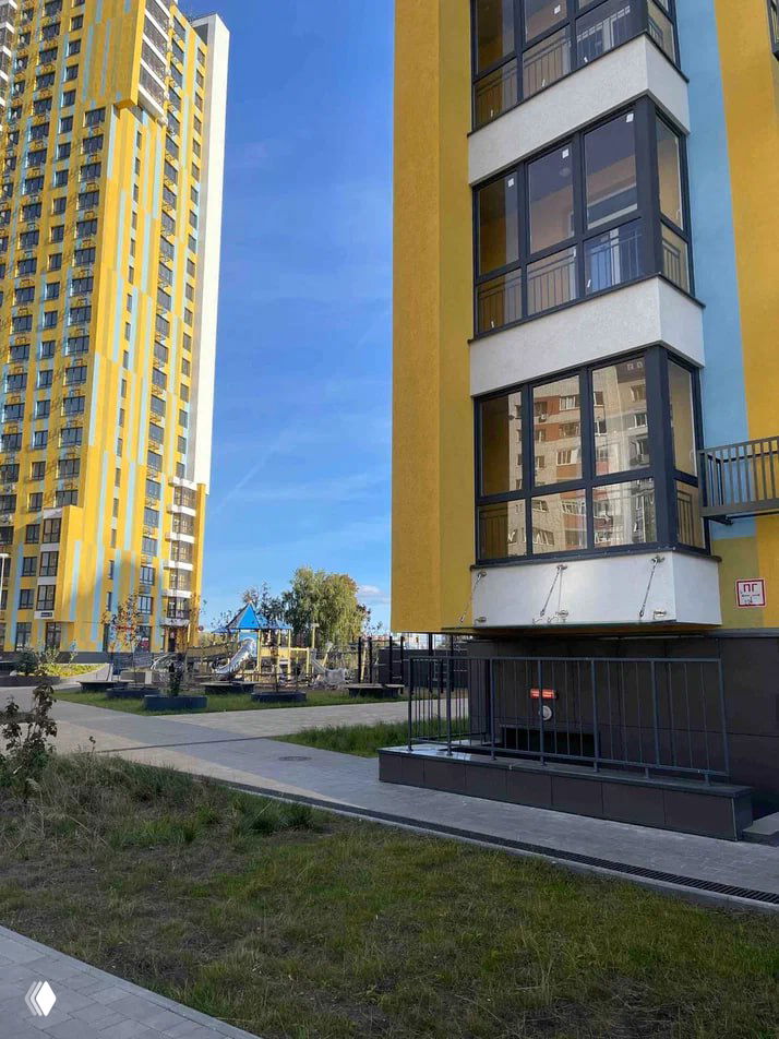 Ближайшая зона у входа в корпус ЖК «Янтарь»: детская площадка, тротуар и часть подвала с видимым ограждением паркинга