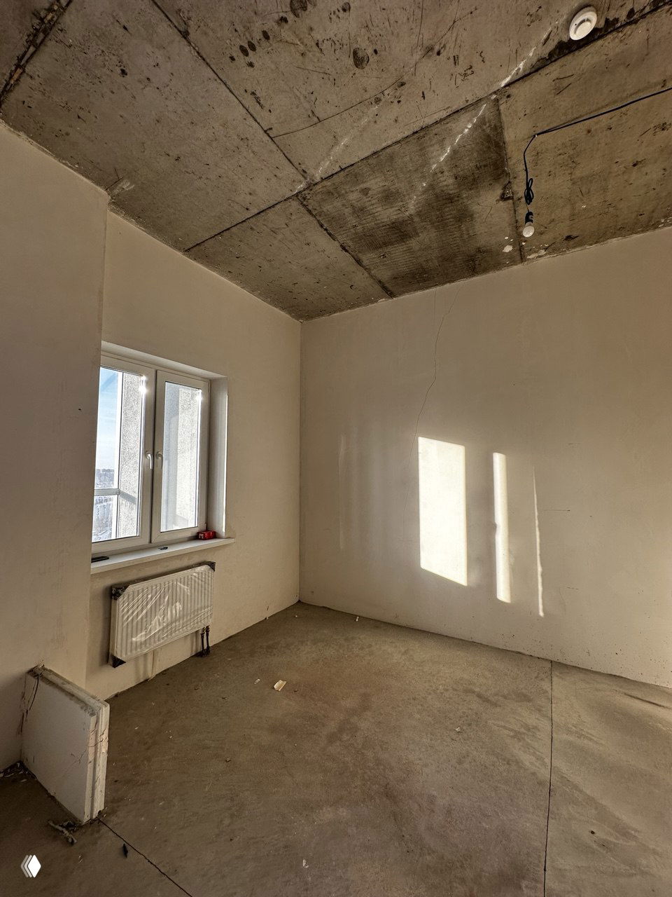 Пустая жилая комната без отделки: бетонный потолок, радиатор под окном, оштукатуренные стены и черновой пол — варианты свободной планировки.