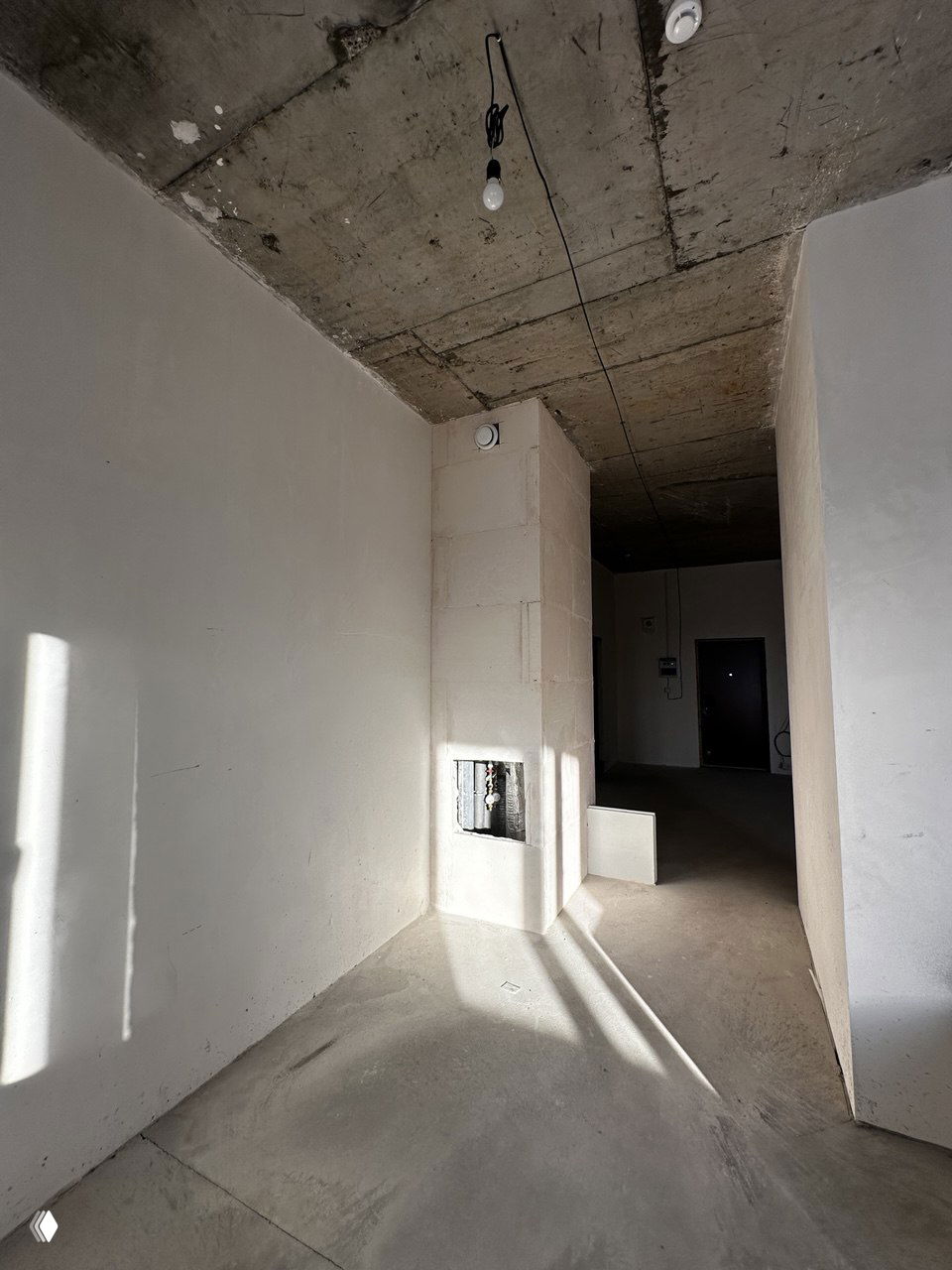 Прихожая и коридор в квартире от застройщика: необработанные стены, элементы инженерных узлов, висящая лампочка и входная дверь на фоне.