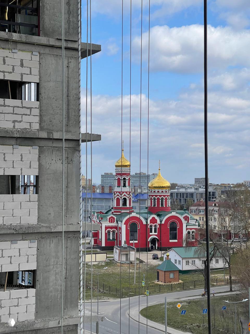 Вид из окна стройплощадки на красную церковь с золотыми куполами: в кадре дорожная развязка, городские здания и вертикальные монтажные тросы на переднем плане.