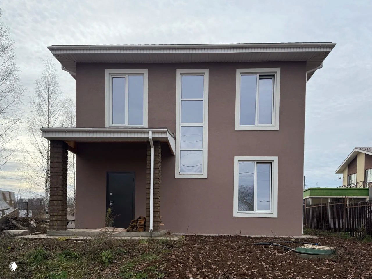 Фасад дома со стороны крыльца: два этажа, высокие окна и навес над входной дверью, участок с земляными разработками на переднем плане.