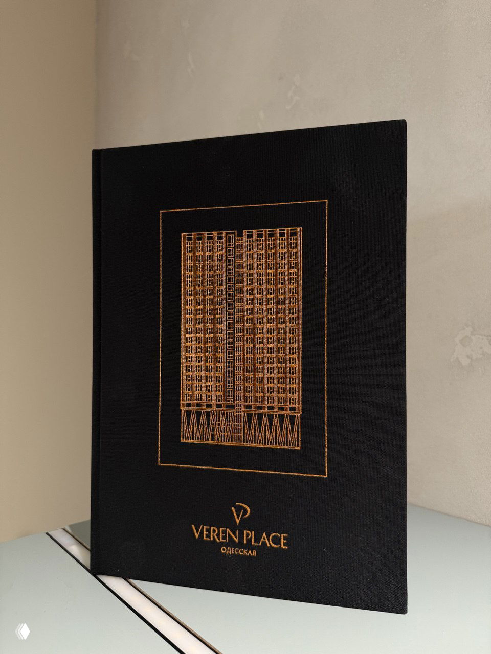 Презентационная папка «Veren Place Одесская» чёрного цвета с тиснёным логотипом и схематическим изображением фасада жилого комплекса на обложке.