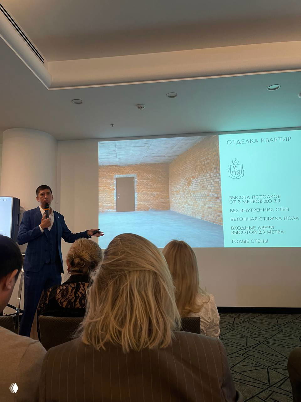 Спикер на презентации показывает слайд с информацией об отделке квартир и параметрах потолков (3–3,3 м), аудитория слушает доклад.