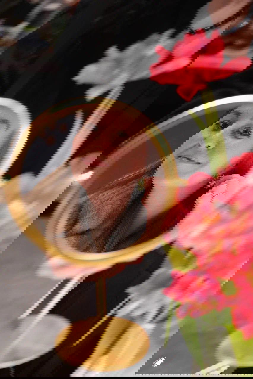 Женщина смотрит в настольное зеркало, примеряя серьгу; на переднем плане размытые яркие цветы, тёплый кадр с маркета.