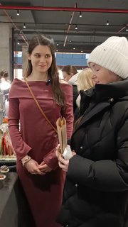 Две женщины на маркете: одна держит растение в горшке, другая в тёплой куртке и белой шапке рассматривает товар.