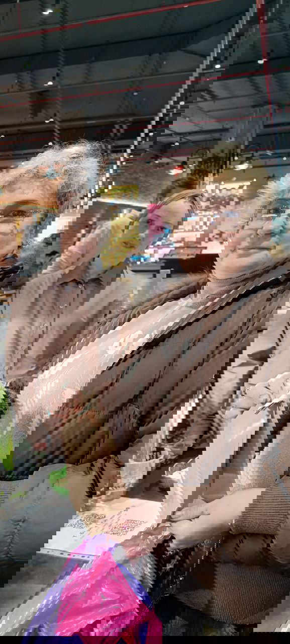 Две посетительницы у прилавка цветочного маркета: одна держит бумажный пакет, на фоне стойка с растениями и торговая зона.