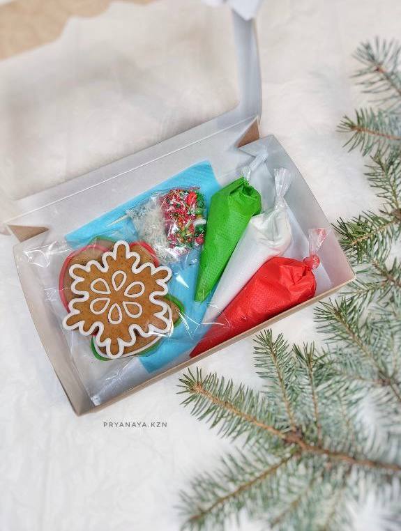 Подарочная коробка с украшенным пряником и тюбиками цветной глазури в праздничной упаковке рядом с еловой веткой.