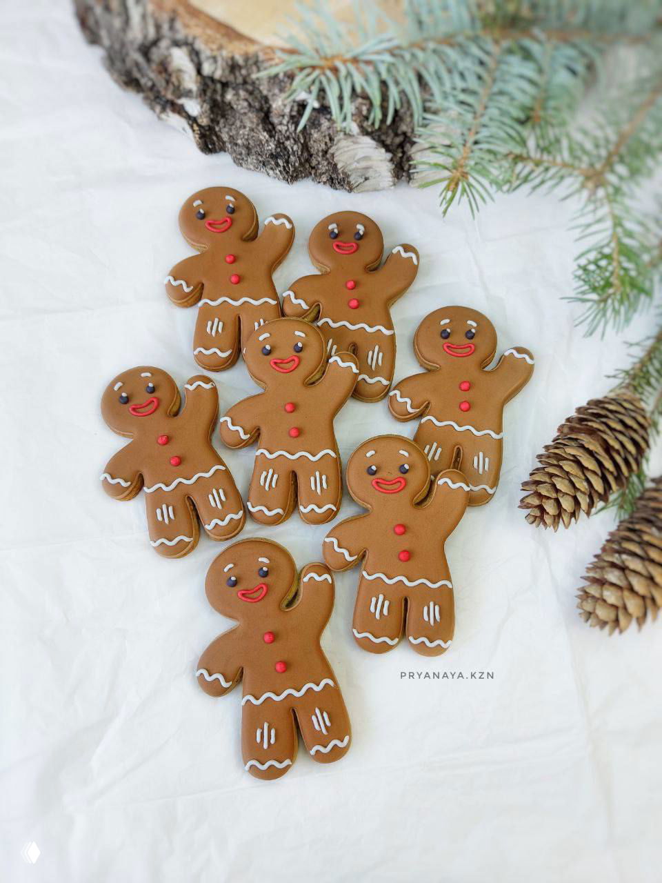 Несколько имбирных пряников в виде человечков, украшенных белой и красной глазурью, выложены на светлой ткани рядом с еловой веткой.