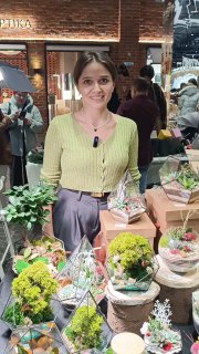 Женщина на маркете рядом со столом, на котором выставлены флорариумы и мосариумы — компактные декоративные композиции с мхом и мелкими растениями.