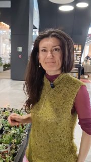 Портрет Диляры — участницы маркета: улыбается и держит комнатное растение, на заднем плане торговое пространство и посетители.