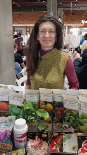 Женщина-продавец за прилавком на цветочном маркете в Казани, на столе пакеты семян, луковичные растения в горшках и банка удобрений.