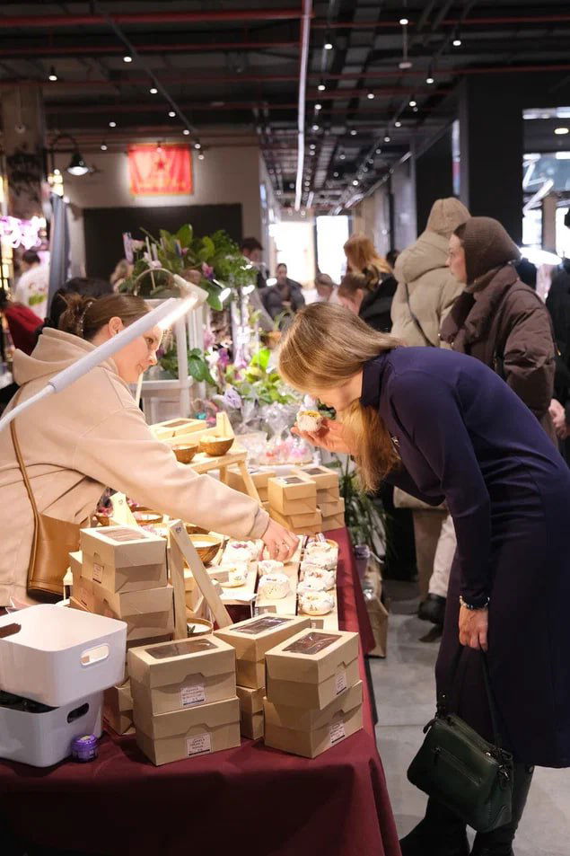 Покупатели изучают хендмейд и эко-товары на ярмарочных прилавках в АРТ Центре: коробки с товарами, продавцы и заинтересованные посетители.