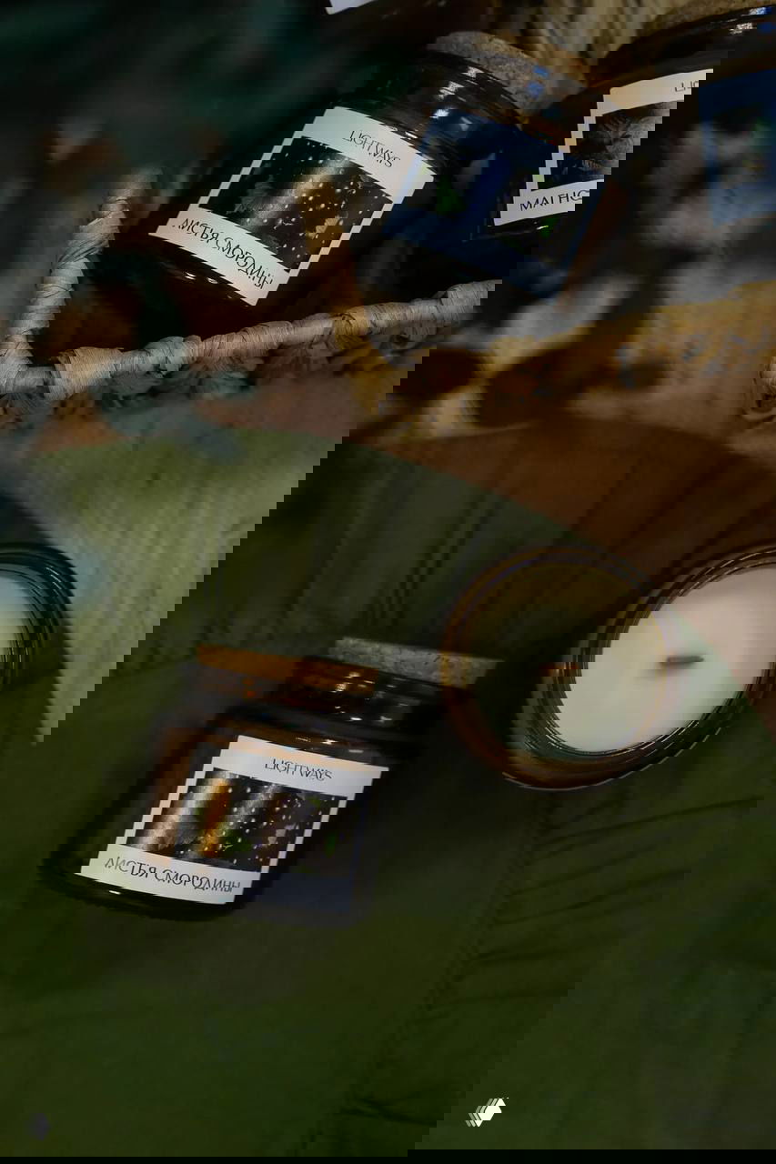 Небольшие стеклянные баночки со свечами и этикетками LIGHT WAYS «ЛИСТЬЯ СМОРОДИНЫ» в корзине и на ткани, крупный план продукта.