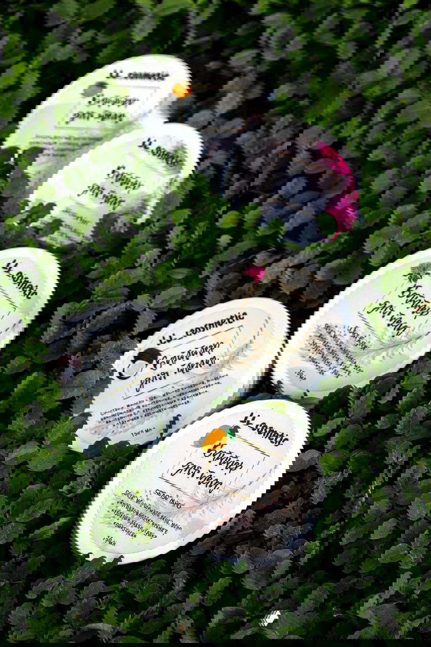 Круглые баночки бальзама и баттера LL_cosmetic, расположенные на зелёной траве (клевер), фотокомпозиция для презентации продукта.