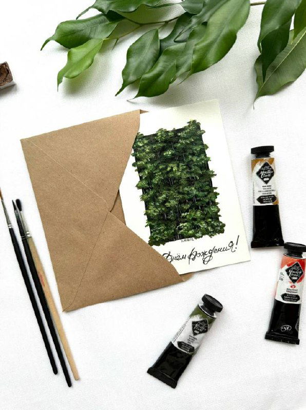 Фотография рабочей сцены: открытка с изображением зелёного леса в крафтовом конверте, рядом кисти, тюбики красок и декоративные листья.