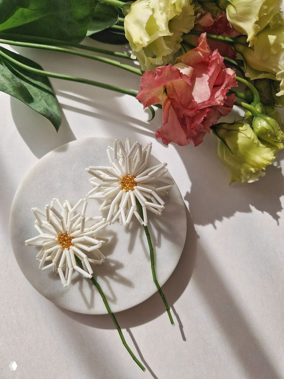 Белые бисерные ромашки в виде серьг лежат на круглой мраморной подставке рядом с живыми цветами — ручная бижутерия с флористическим мотивом.