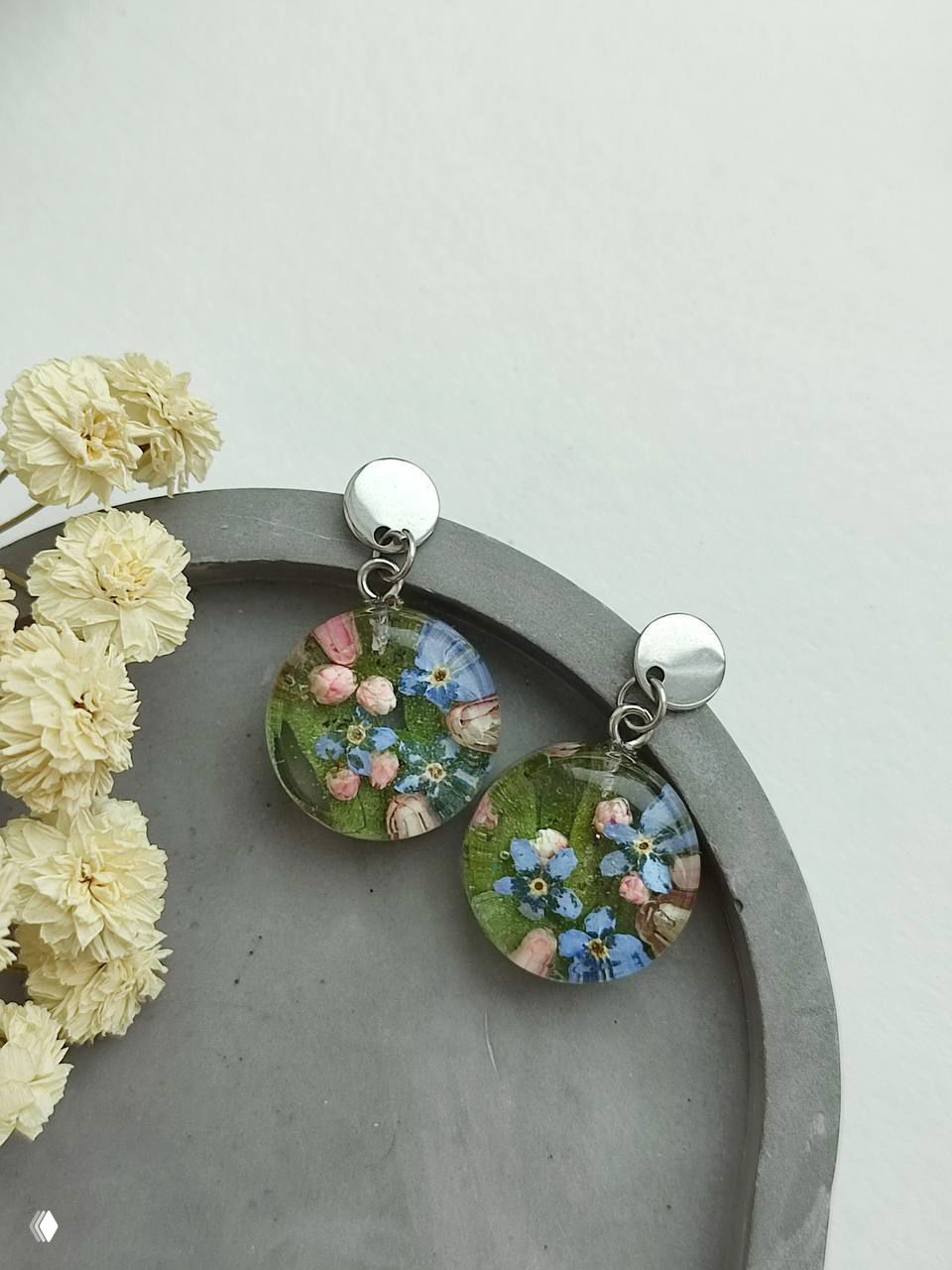 Круглые прозрачные серьги с запечатанными в эпоксидной смоле полевыми цветами на серой подложке — украшения с сухоцветами и смолой.