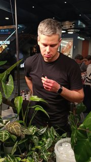 Кадр интервью: мужчина рассматривает растение среди витрины с декоративными листьями; в кадре видна торговая суета и стакан с напитком.