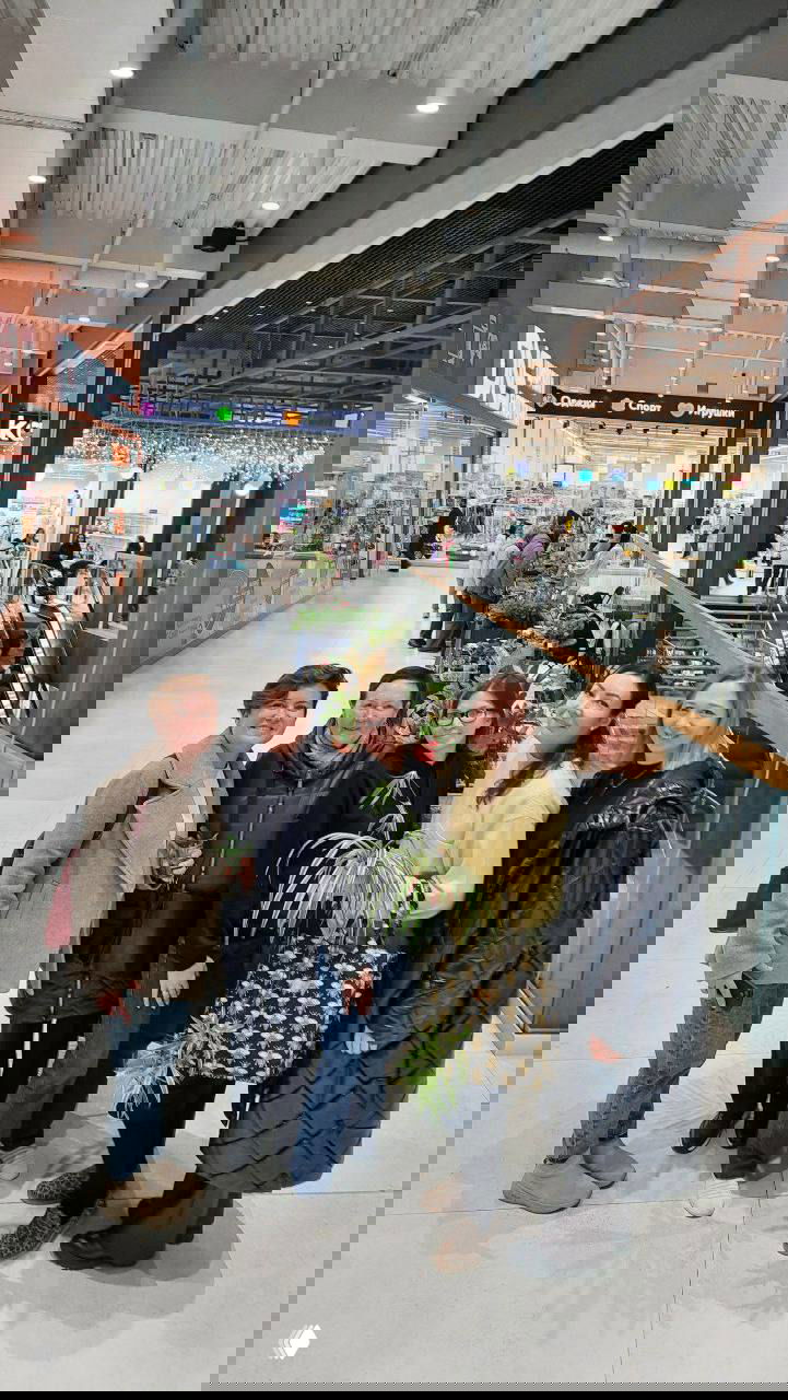 Группа организаторов и участниц маркета в торговом центре с растениями на фоне эскалатора и торговых рядов, атмосферный кадр мероприятия.