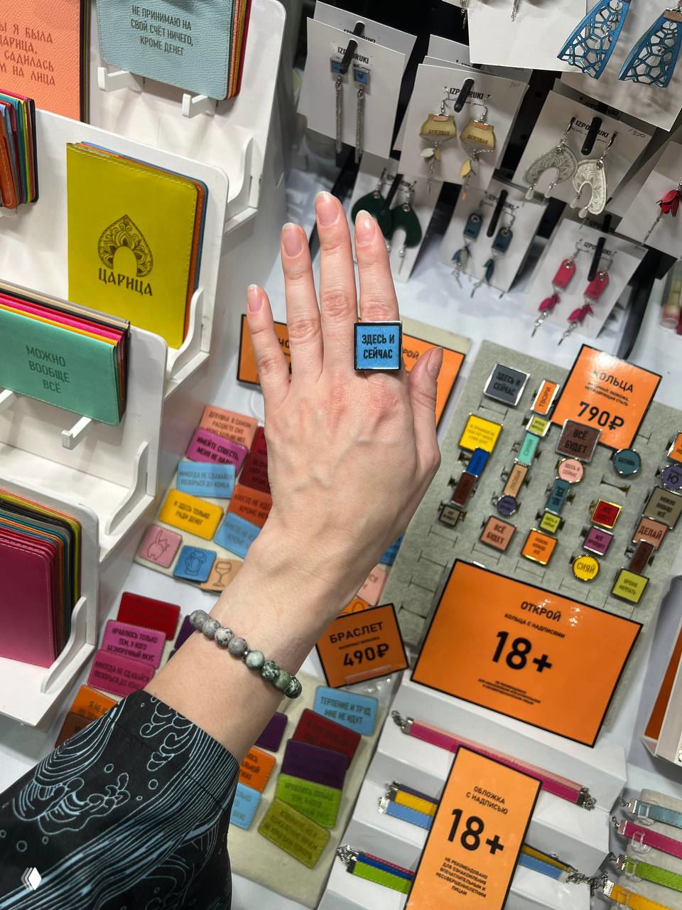 Рука с кольцом показывает изделие над столом с яркими ценниками, значками и аксессуарами на выставочном прилавке маркета.