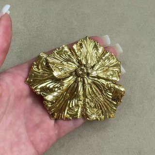 Крупный план золотой броши в форме цветка в ладони: рельефная текстура лепестков, блеск и художественная обработка металла.