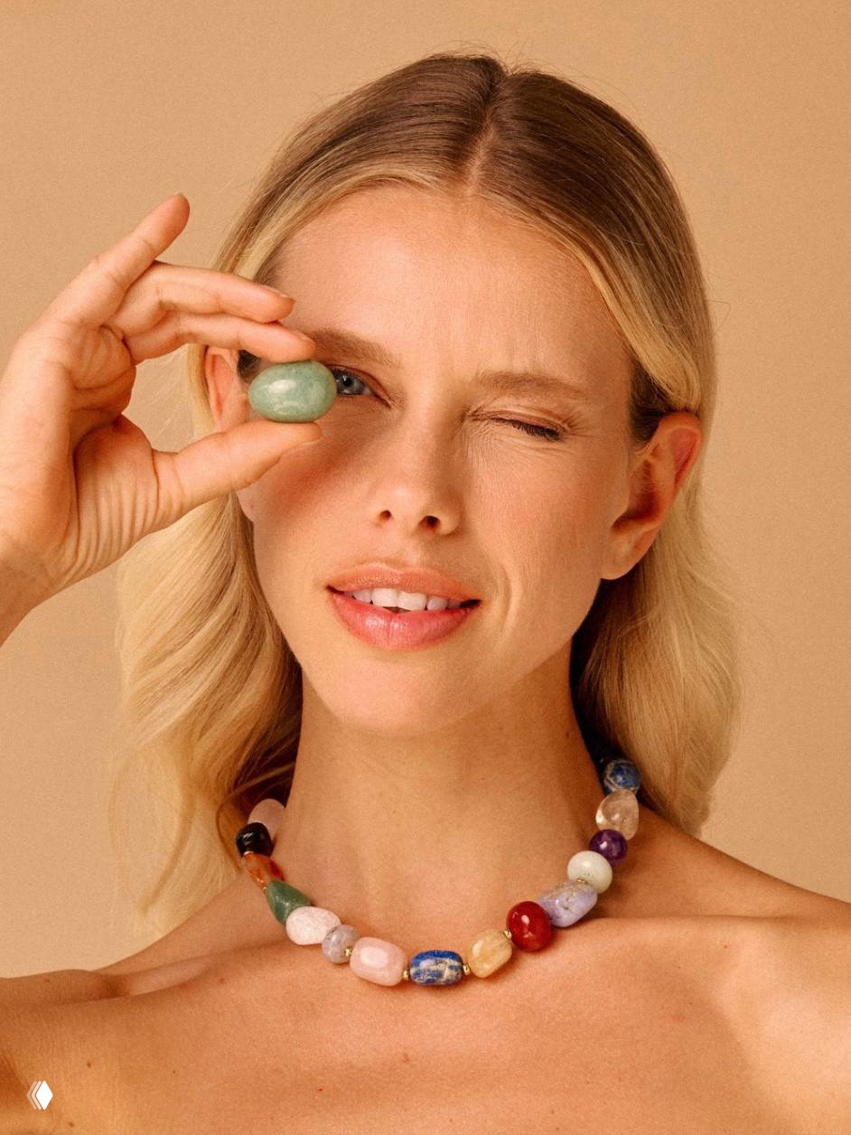 Портрет модели в ожерелье Nature Bijoux из крупных полированных бусин разного цвета; модель держит камень у глаза на светлом фоне.