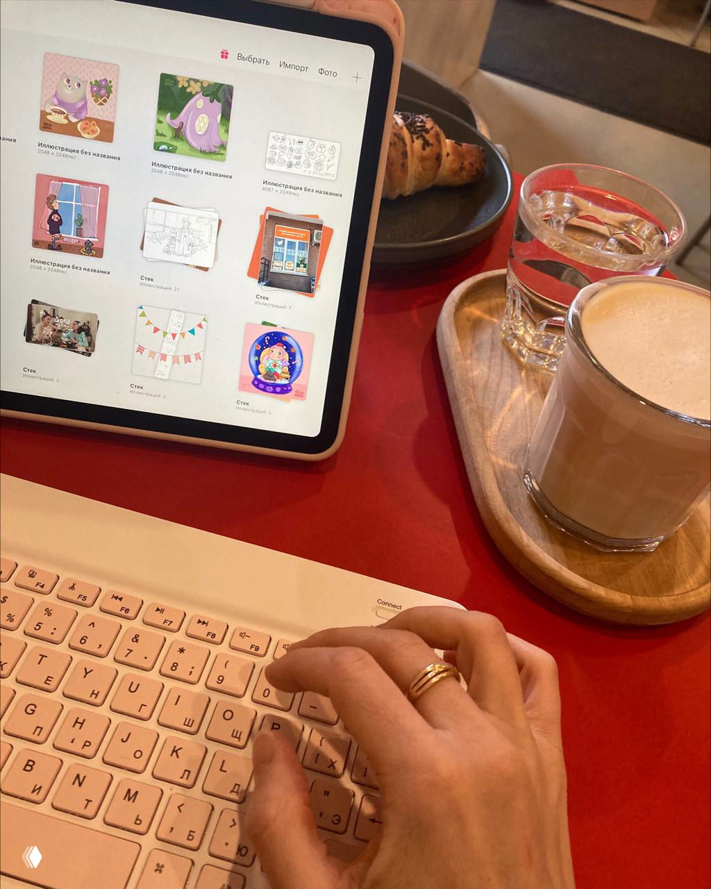 Рабочая сцена в кафе: на столе iPad с эскизами и миниатюрами иллюстраций, беспроводная клавиатура, рогалик, стакан воды и чашка с кофе на подносе — уютное место для работы.