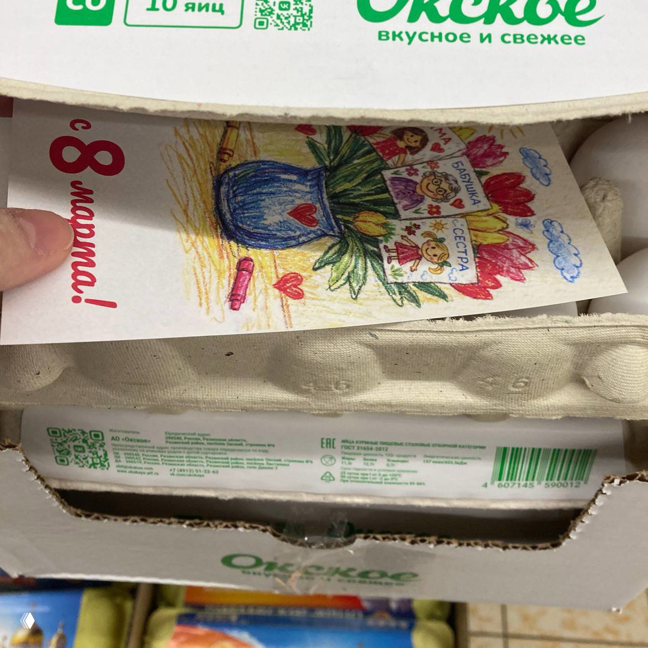 Открытка с цветным детским рисунком (ваза, тюльпаны, надпись «с 8 марта!») частично видна в раскрытой картонной упаковке яиц в магазине.