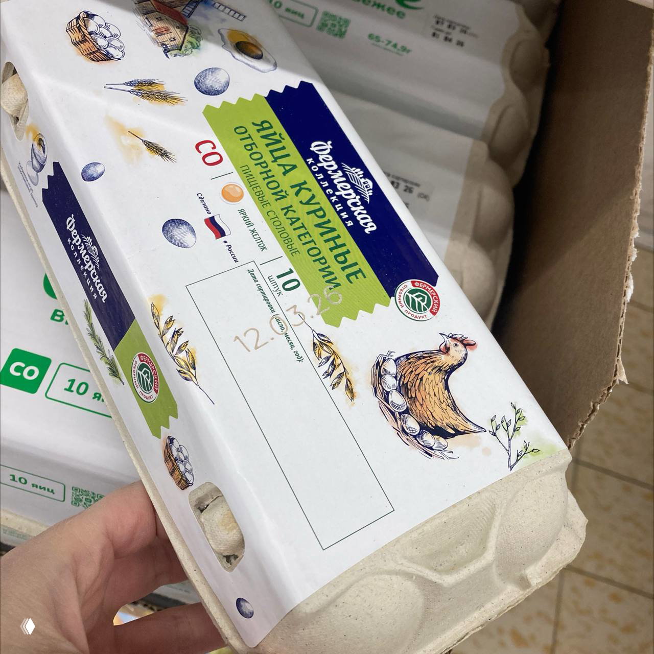 Крупный план картонной упаковки яиц с фирменной этикеткой: иллюстрация курицы, пиктограммы и указание категории яйца, упаковки сложены в коробке магазина.