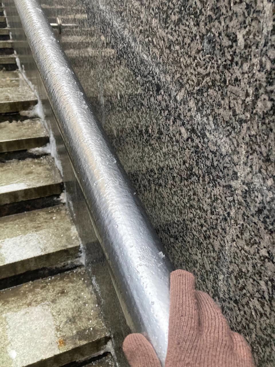 Обледеневший металлический перила лестницы в уличном переходе; рука в перчатке держится за холодный поручень в дождливую погоду