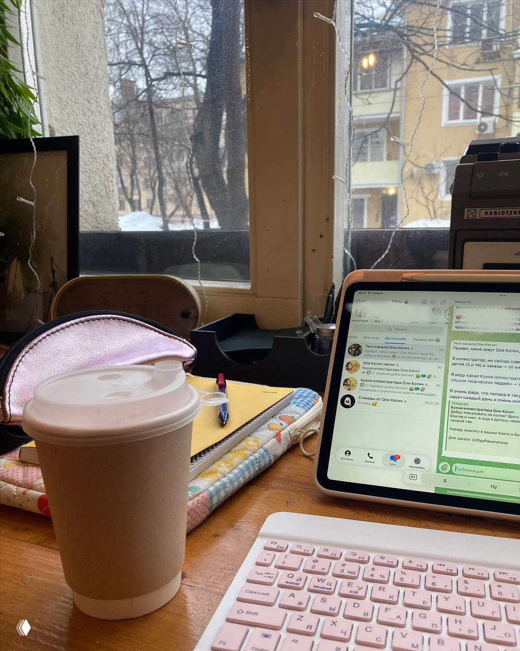 Кофе в бумажном стакане, планшет с открытыми сообщениями, блокнот и розовая клавиатура на рабочем столе у окна со снегом.