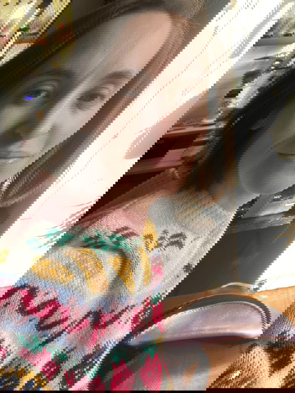 Женщина в свитере с фруктовым узором сидит за столом в студии; на заднем плане кофемашина и предметы рабочего интерьера.