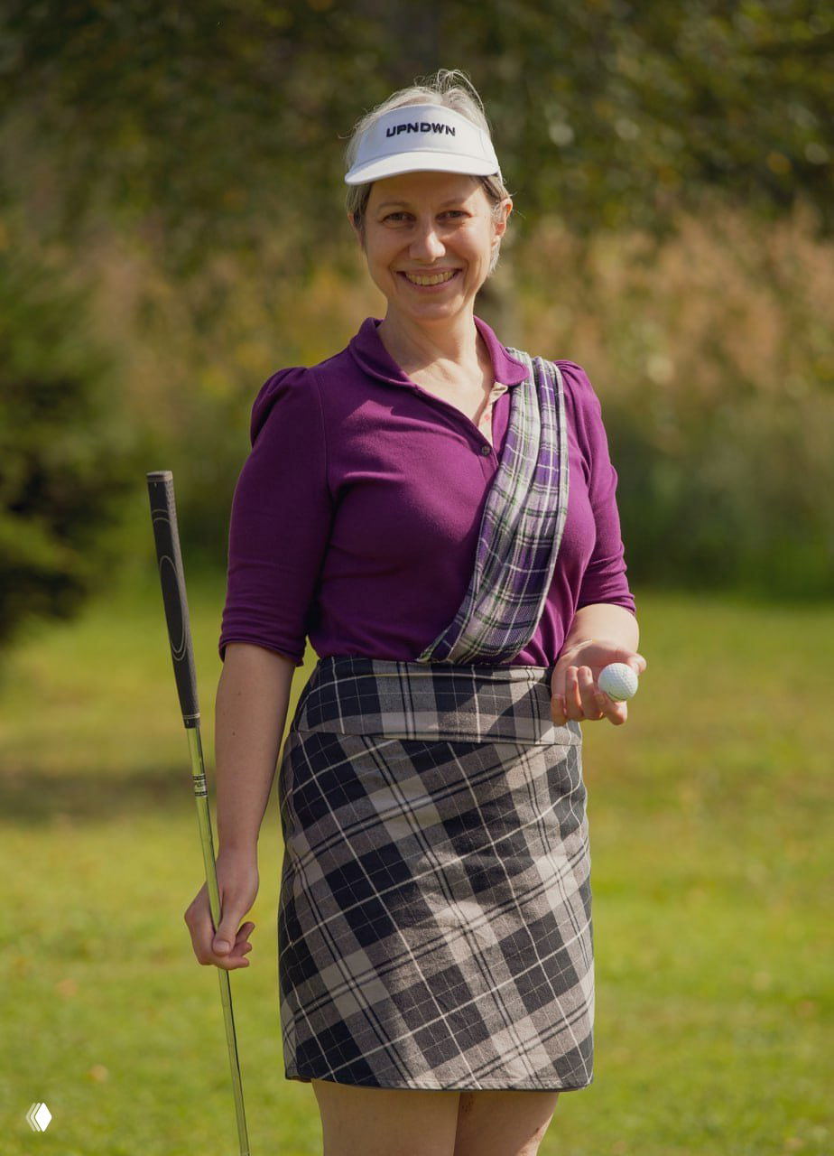 Женщина в гольф-экипировке на поле: клюшка в руке и мяч, спокойная улыбка, природный фон — символ спортивного подхода.