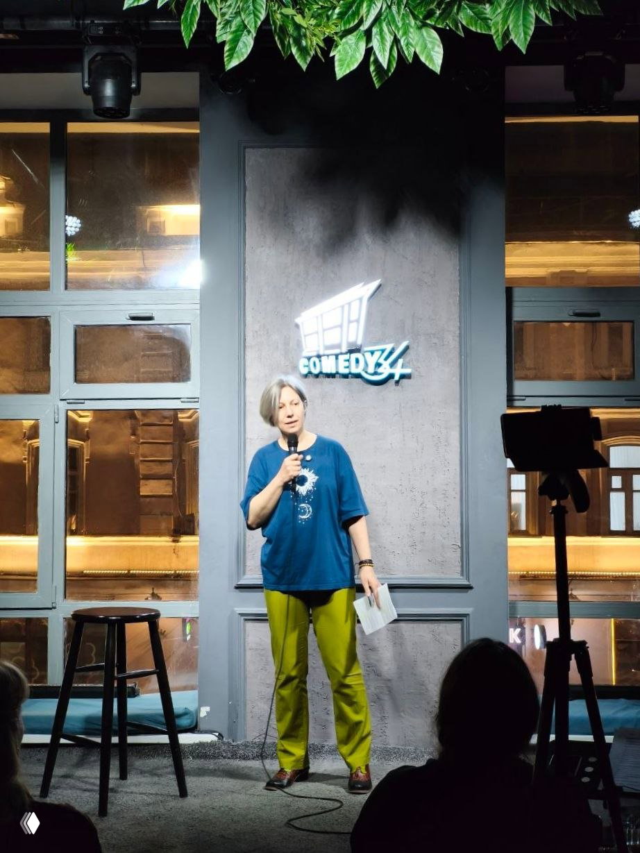 Автор выступления на сцене Comedy 34 с микрофоном и листом заметок, в синей футболке и салатовых брюках; свет сцены и окно с видом на улицу за сценой.