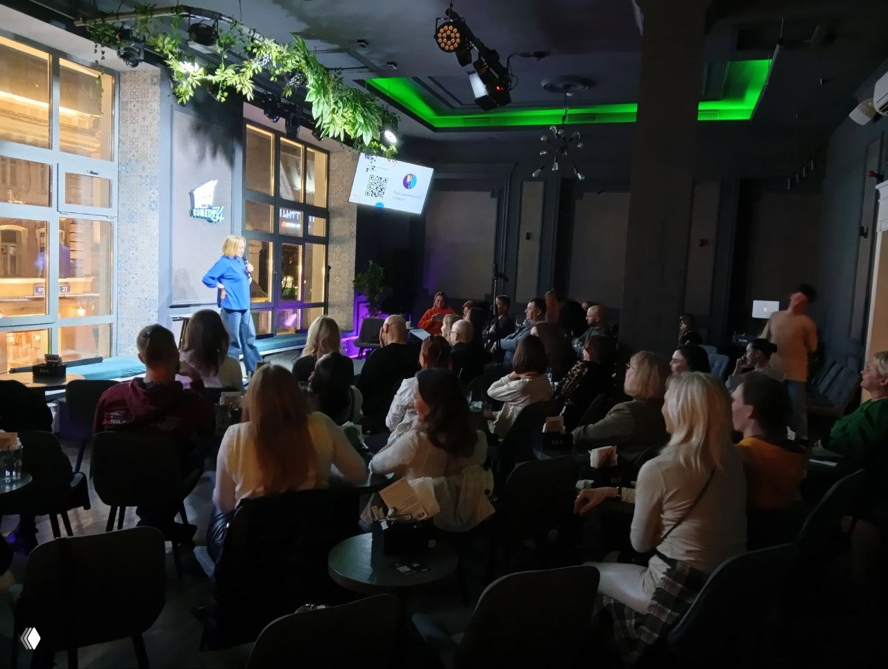 Широкий кадр зала Comedy 34: аудитория за небольшими столиками смотрит на сцену, экран над сценой, зелёные декорации и тёплая сценическая подсветка.