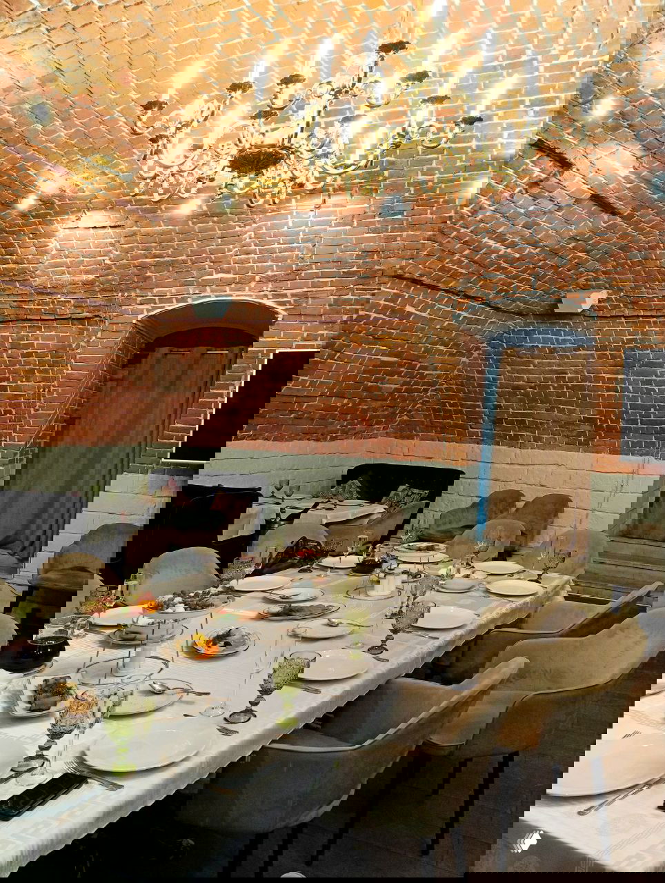 Большой столи украшен белой скатертью, закусками и зелёными бокалами в кирпичном подвале с люстрой — подготовка к обеду после литературной встречи.