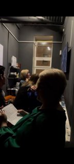 Узкая закулисная комната перед выходом на сцену: несколько участников готовятся, читают заметки и ждут своей очереди в приглушённом свете.