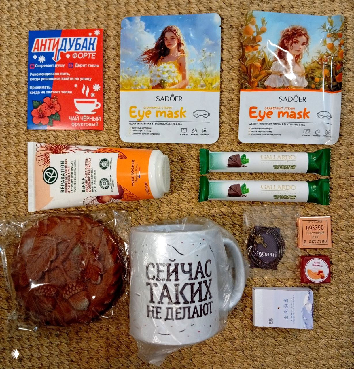 Фото содержимого набора, разложенного на фоне: маски для глаз, шоколадки, крем, пряник, кружка с надписью и небольшие саше/карточки.