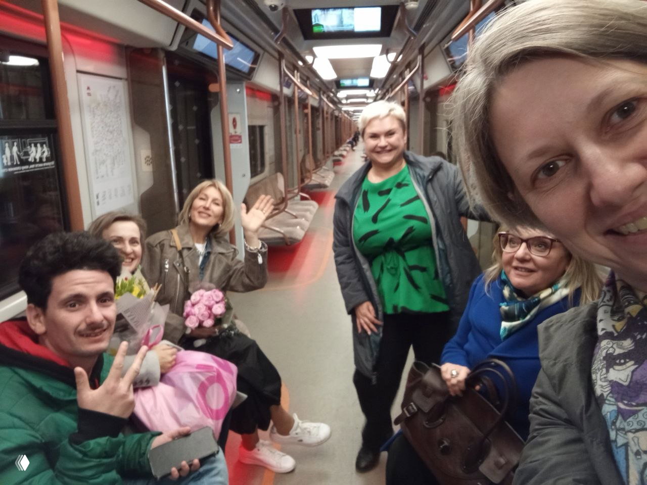 Групповое селфи в вагоне метро после выступления: несколько людей с букетами и сумками улыбаются в камеру, праздничное настроение и возвращение домой командой.
