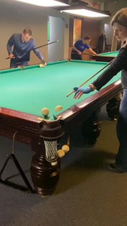 Игроки за бильярдным столом в закрытом помещении: мужчина и женщина готовятся к удару, видно зелёное сукно и разноцветные шары