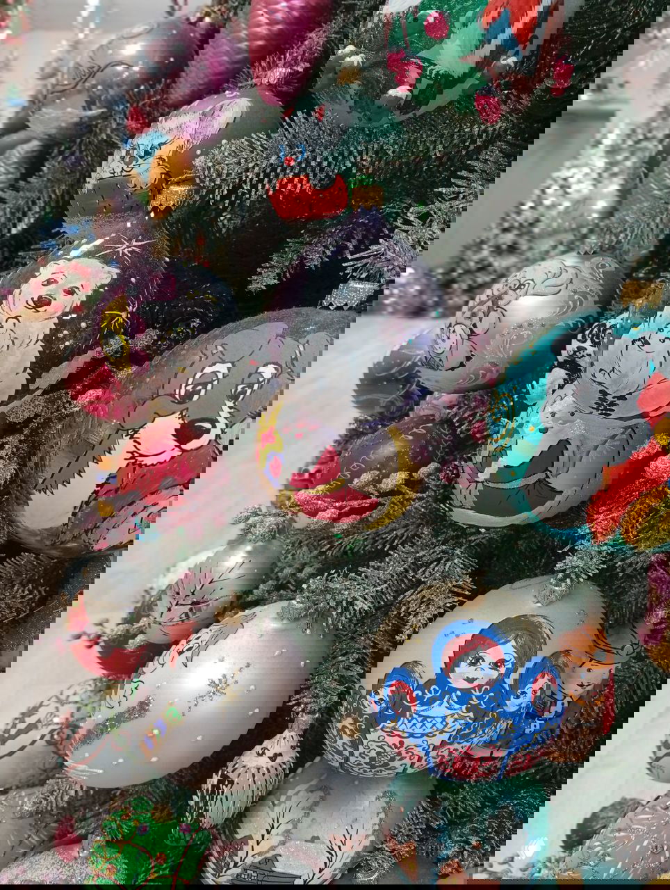 Крупный план расписных ёлочных шаров с матрёшками, белым медвежонком и другими русскими мотивами на украшенной живой ёлке в витрине.