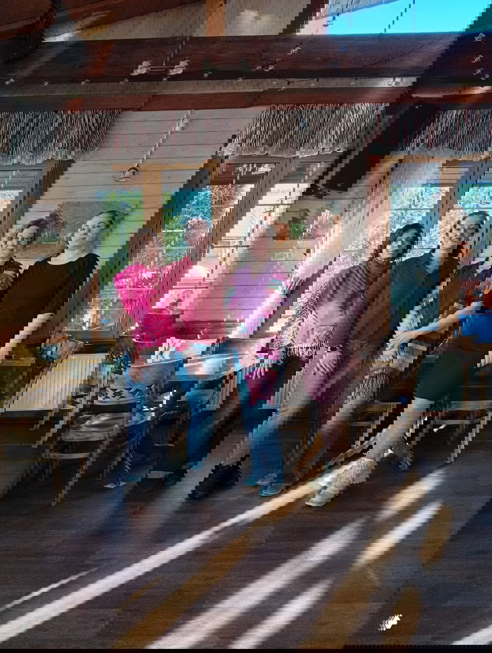 Четверо участниц выезда стоят в тёплом деревянном зале с солнечными бликами на полу; дружеская сцена в бордовых оттенках интерьера.