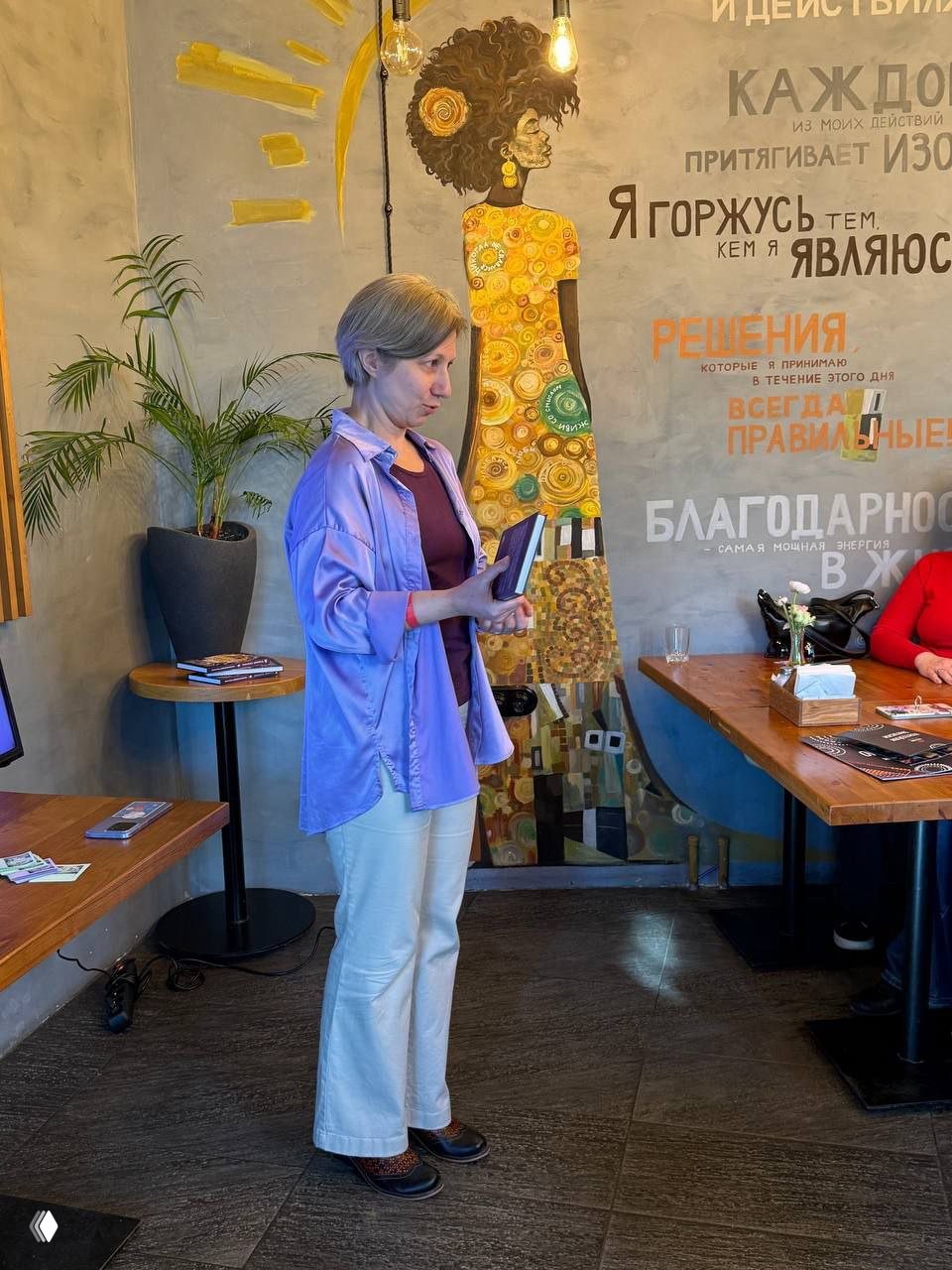 Автор рассказывает публике о книге на фоне стильной настенной росписи, держит книгу в руках и делится выдержками с аудиторием.
