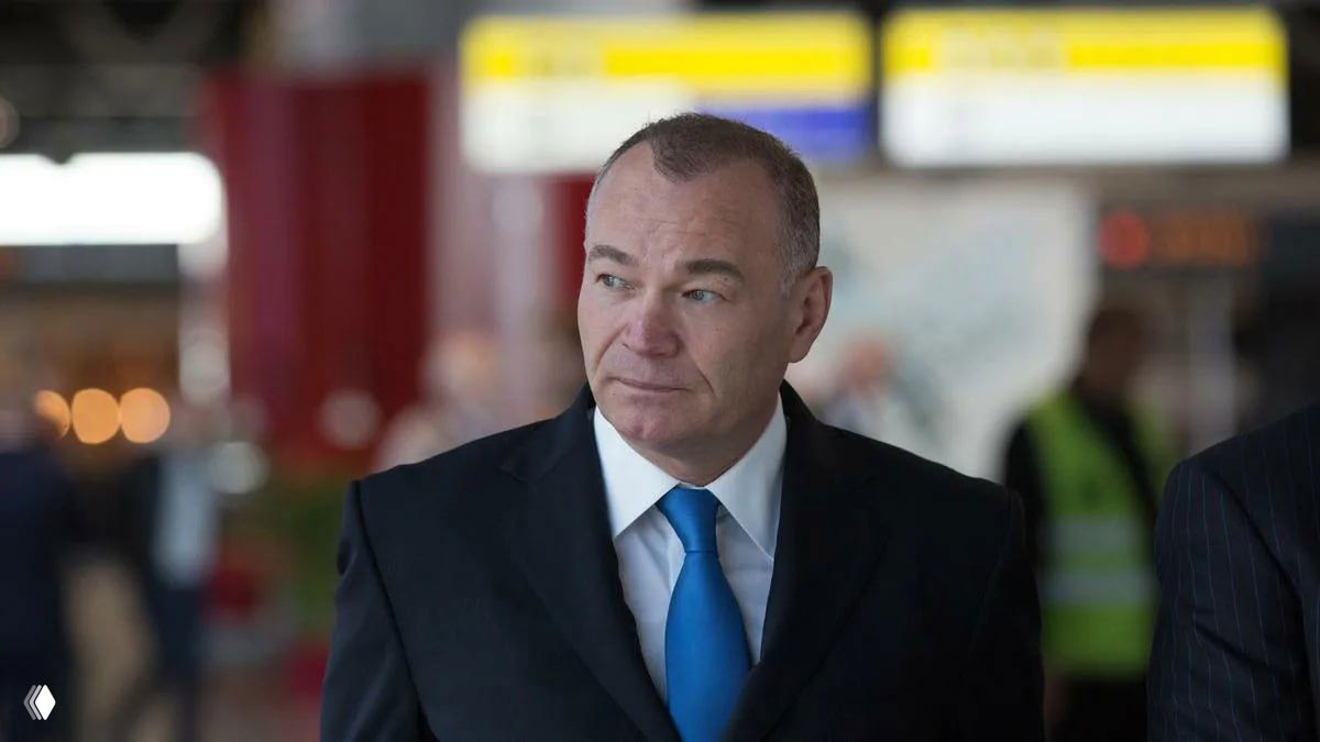 Мужчина в деловом костюме на фоне зала аэропорта с табло и пассажирами; иллюстрация к теме продажи аэропорта.