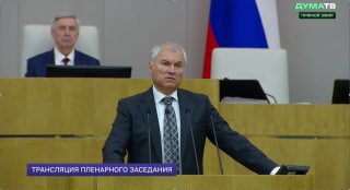 Оратор за трибуной в зале пленарных заседаний Госдумы: выступление перед депутатами, на заднем плане виден российский флаг и надпись трансляции.