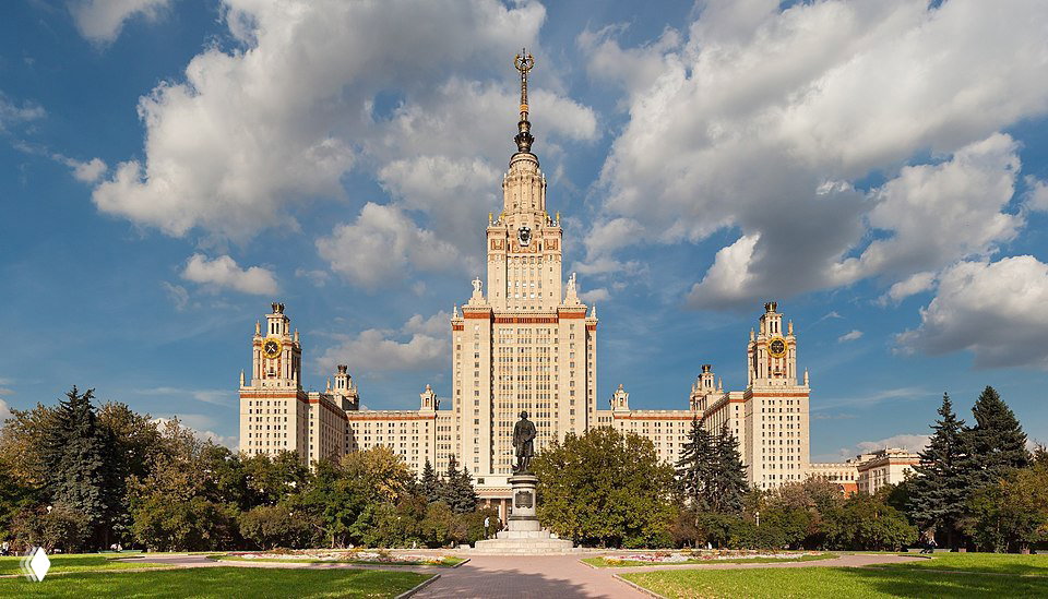 Фасад главного корпуса МГУ им. Ломоносова на фоне голубого неба и облаков, вид спереди с памятником и партерной аллеей и зеленью