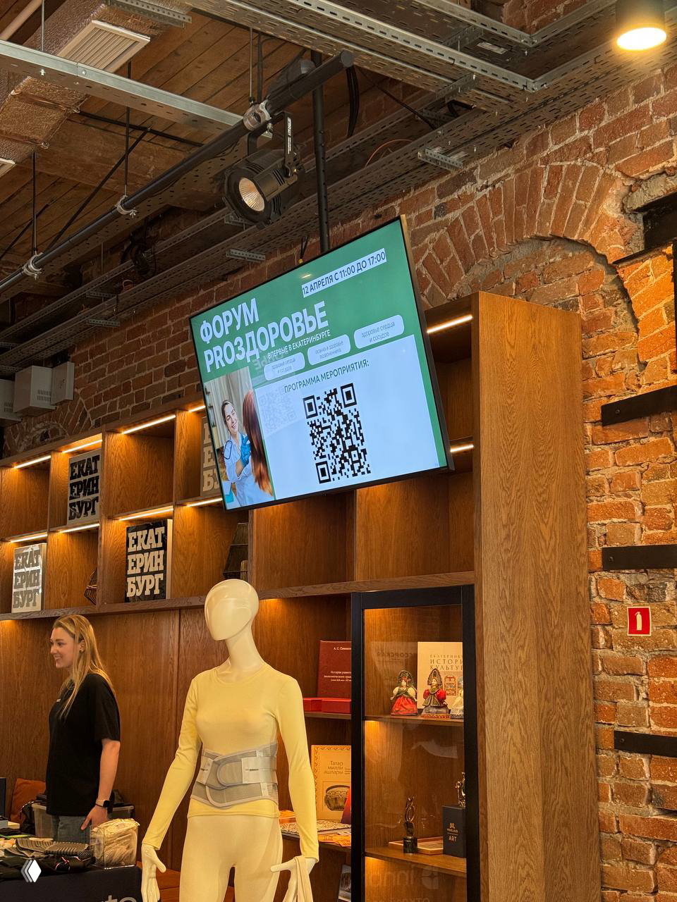 Электронный экран с объявлением «Форум PROЗдоровье», манекен и стенды на фоне кирпичных стен креативного кластера Домна.