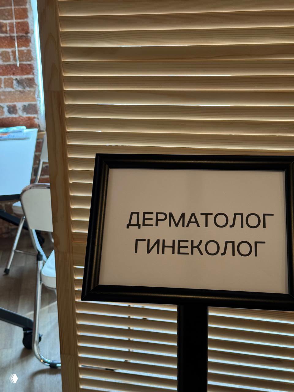 Фотография таблички «ДЕРМАТОЛОГ ГИНЕКОЛОГ» на подставке перед зоной приёма; деревянная ширма и интерьер помещения видны.