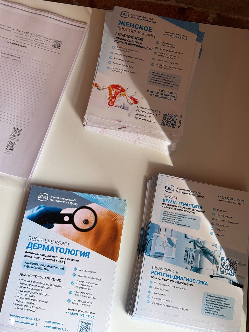 Стопка буклетов и информационных материалов Екатеринбургского Медицинского Центра о дерматологии, гинекологии и диагностике на столе.