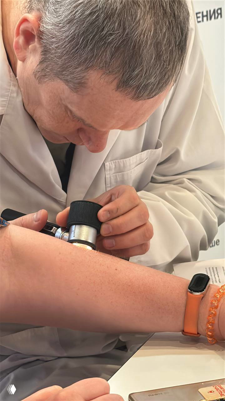 Врач в белом халате осматривает участок кожи пациента с помощью дерматоскопа во время приёма на стенде клиники на форуме.